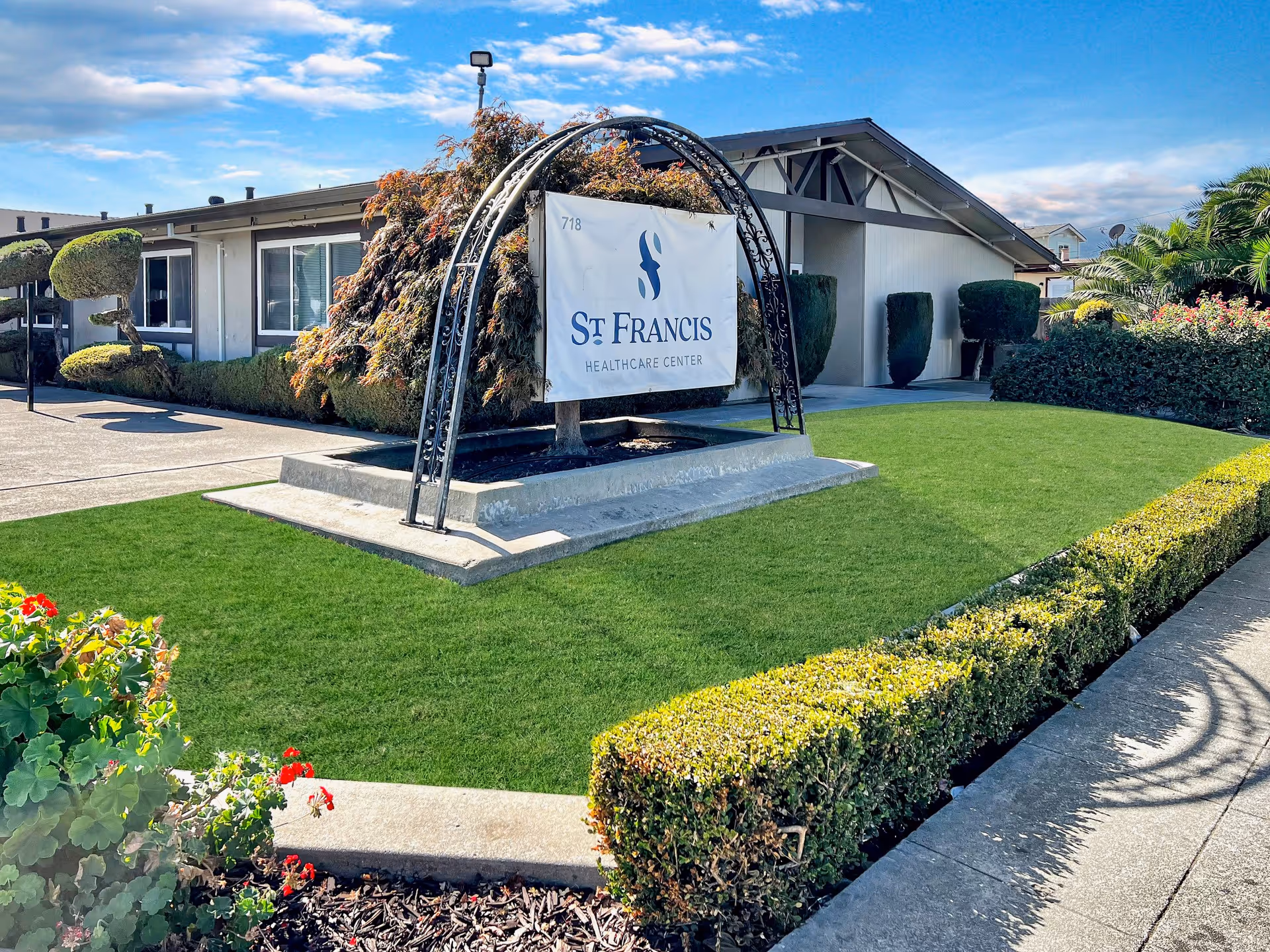 Exterior view of St. Francis Healthcare Center showing a well-maintained lawn, trimmed bushes, a decorative arch with a sign displaying the facility's name, and a single-story building under a blue sky with some clouds.