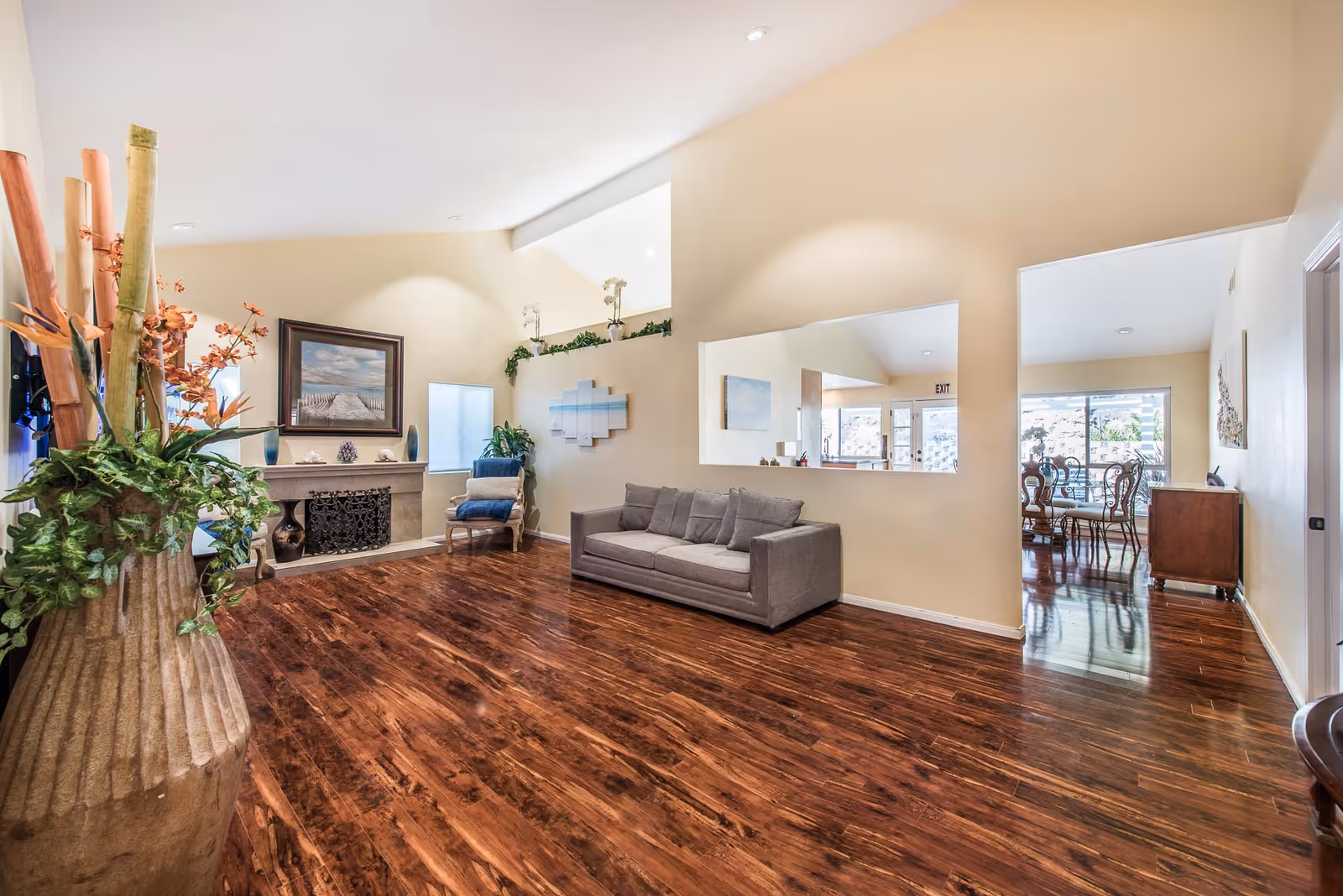 A spacious living room with polished wooden floors, a gray sofa with cushions, a decorative fireplace with a framed painting above it, a blue armchair, and a large vase with tall bamboo and flowers. The walls are painted beige, and there is a large rectangular mirror on one wall. Through an open doorway, a dining area with a wooden dining table and chairs is visible.
