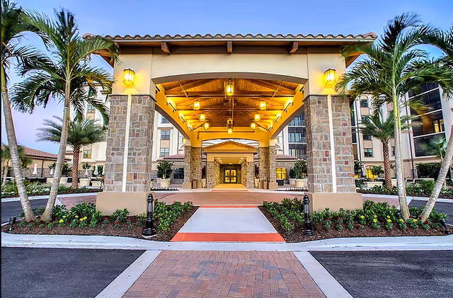 Covered porte-cochère entrance of a multi-story Mediterranean-style building illuminated by hanging lanterns, flanked by palm trees and landscaping.