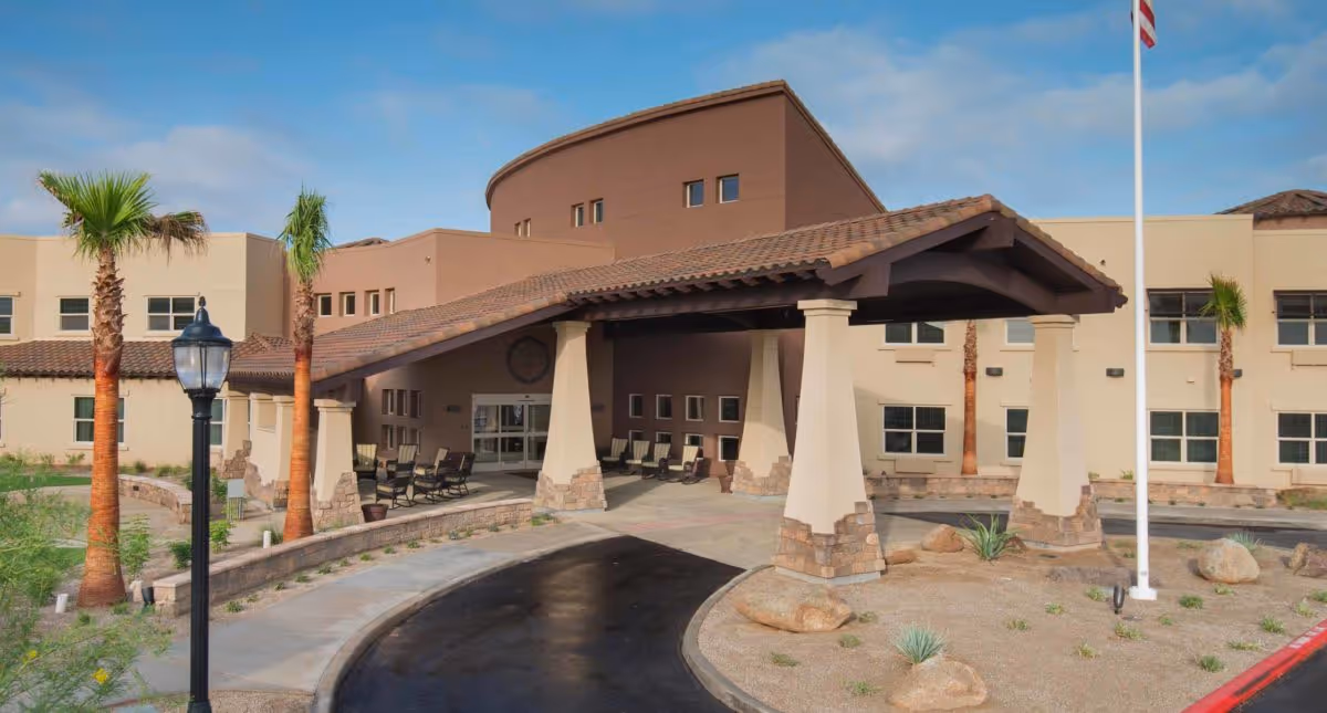 Front entrance and porte-cochère of a beige and brown assisted living building with palm trees and a circular driveway.
