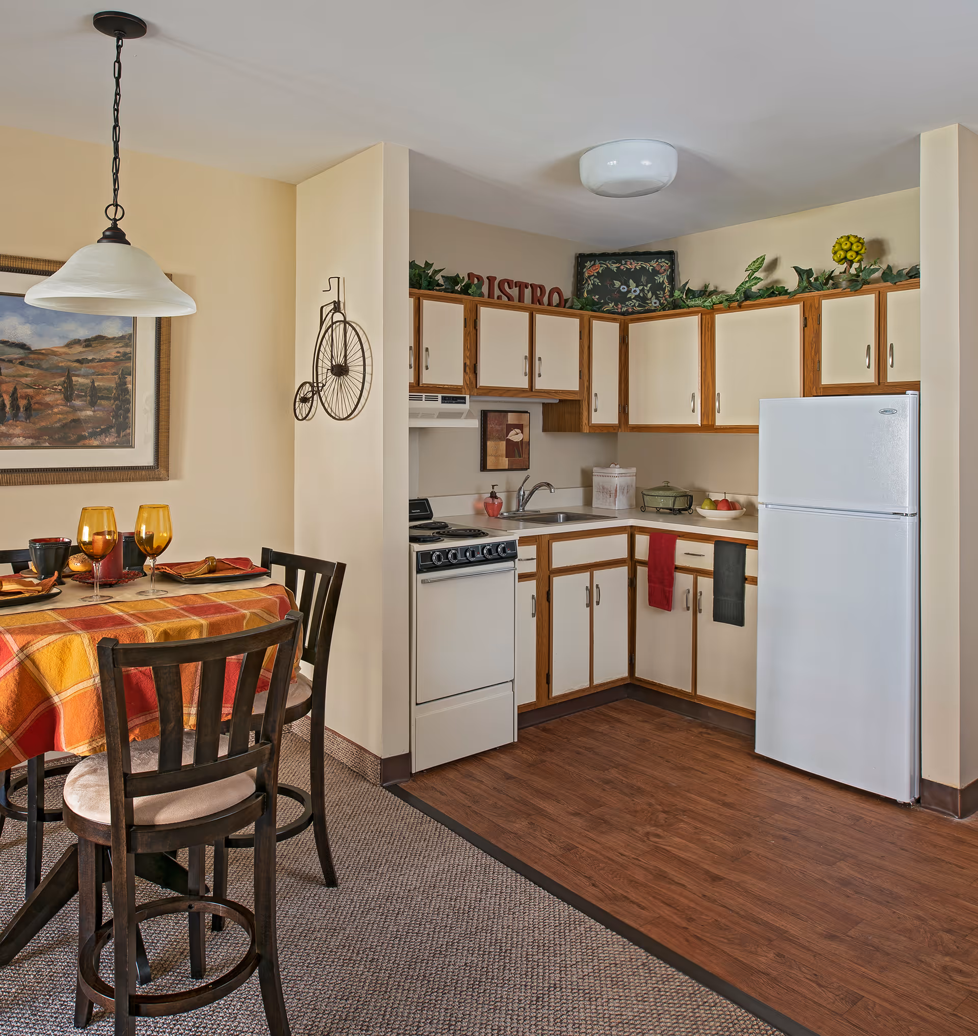 Small open kitchen with white cabinets, refrigerator and stove next to a dining table set with a plaid tablecloth and glasses.