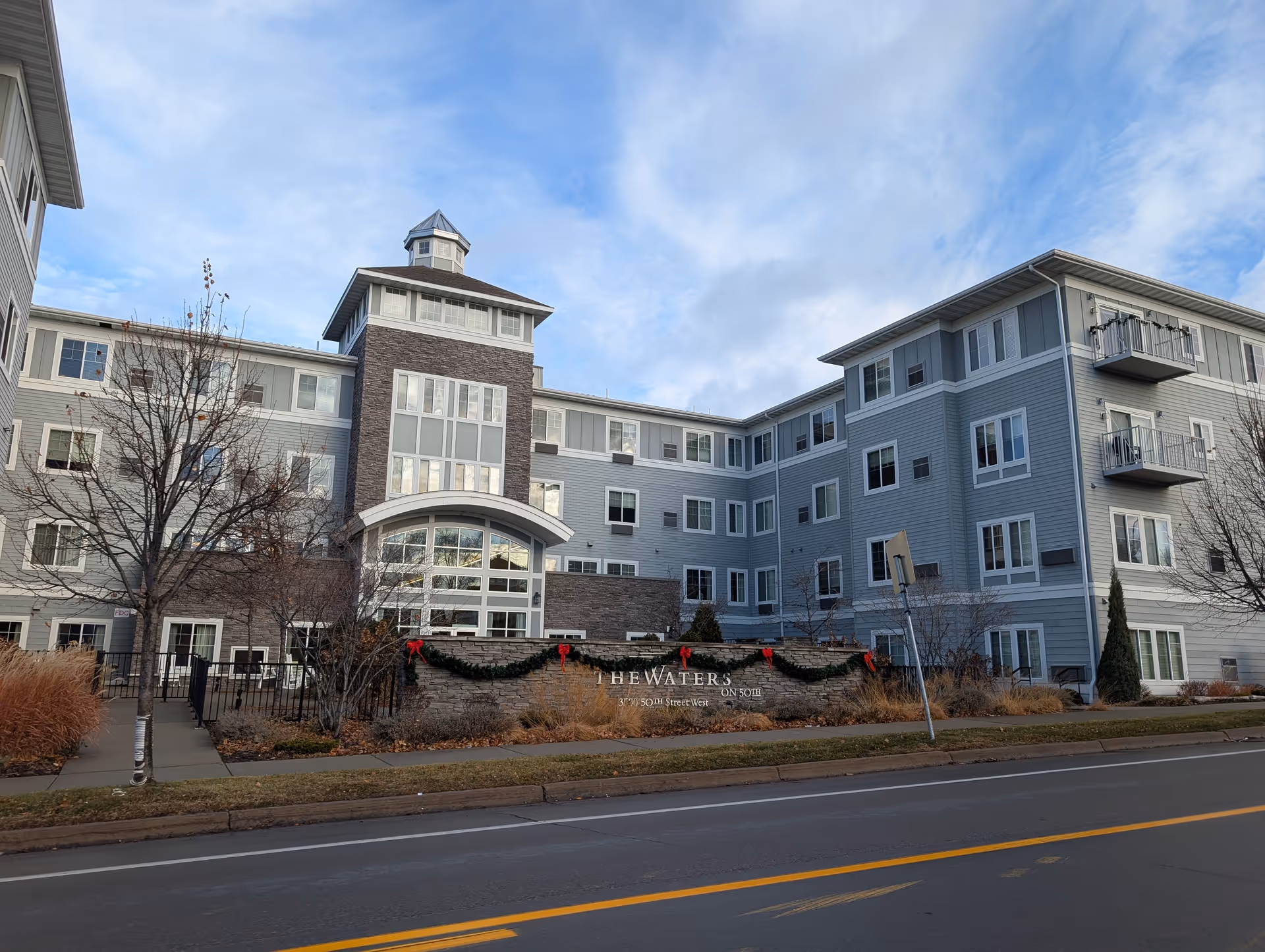 Front exterior of a multi-story senior living building named "The Waters" with landscaped grounds and holiday garlands at the entrance.