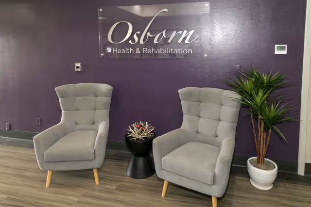 Two gray upholstered armchairs with wooden legs positioned on either side of a small black side table with a decorative arrangement, set against a purple wall with a clear sign that reads 'Osborn Health & Rehabilitation'. A potted green plant is placed to the right of the chairs.