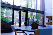 Interior view of a common area in a senior living facility with large glass doors leading to an outdoor patio. The room has a round table with chairs and a floral centerpiece. Outside, there are patio tables with umbrellas surrounded by greenery and trees.