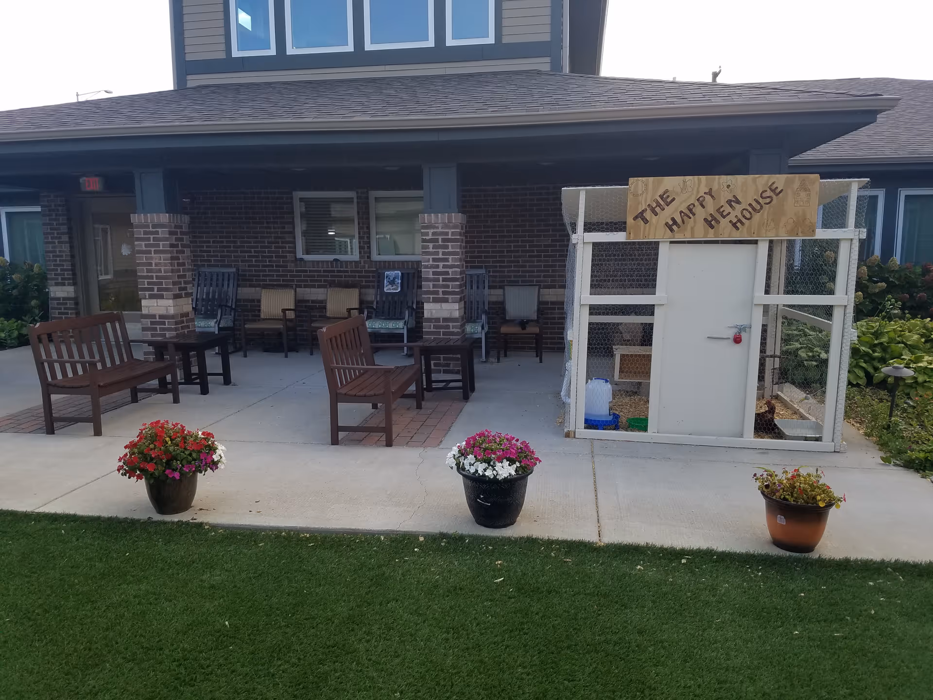 Outdoor patio area of Porter Place with several wooden benches and chairs arranged under a covered porch. In front of the porch, there are three flower pots with colorful flowers placed on the concrete walkway. To the right, there is a small white chicken coop labeled 'The Happy Hen House' with a wooden sign on top.