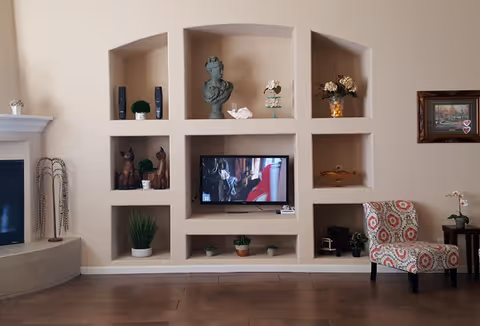 A living room wall with built-in shelves displaying decorative items such as plants, sculptures, and figurines. A flat-screen TV is mounted in the center shelf showing a scene from a movie or show. To the right, there is a patterned upholstered chair next to a small side table with a flower vase. On the left side, part of a fireplace is visible.