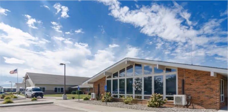 Exterior view of a single-story brick building with large windows under a partly cloudy blue sky. There is a parking lot in front with a white van and an American flag on a flagpole.