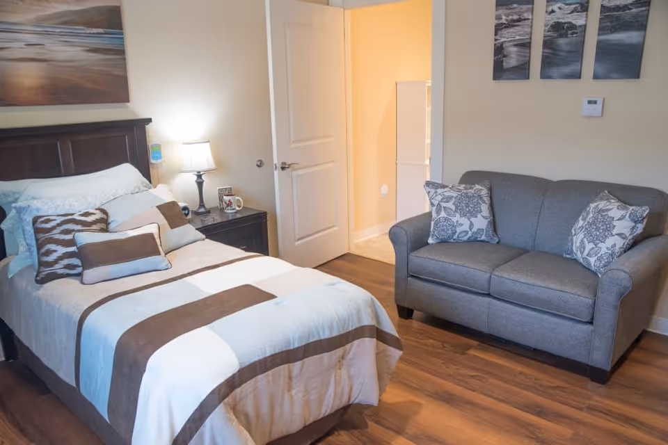 A cozy bedroom featuring a single bed with multiple pillows and a striped bedspread in shades of brown, blue, and white. Next to the bed is a dark wooden nightstand with a lamp, a mug, and a small clock. A gray loveseat with two floral-patterned pillows is positioned against the opposite wall. The room has wooden flooring, light-colored walls, and three framed pictures of ocean scenes above the loveseat. A door is open, revealing a hallway with light-colored walls and flooring.