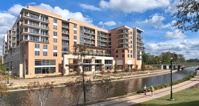 A large multi-story residential building with balconies overlooking a canal. There is a paved walkway along the canal with two people walking. The sky is partly cloudy and there are trees and grass along the walkway.