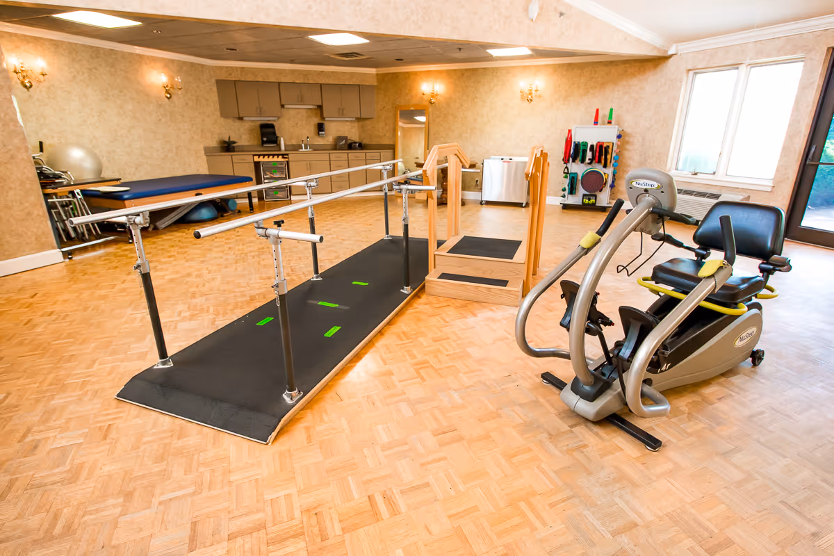 A rehabilitation and wellness room featuring parallel bars for walking exercises, a wooden step platform, a recumbent exercise bike, and various physical therapy equipment in the background. The room has wooden parquet flooring, beige walls, and a window letting in natural light.