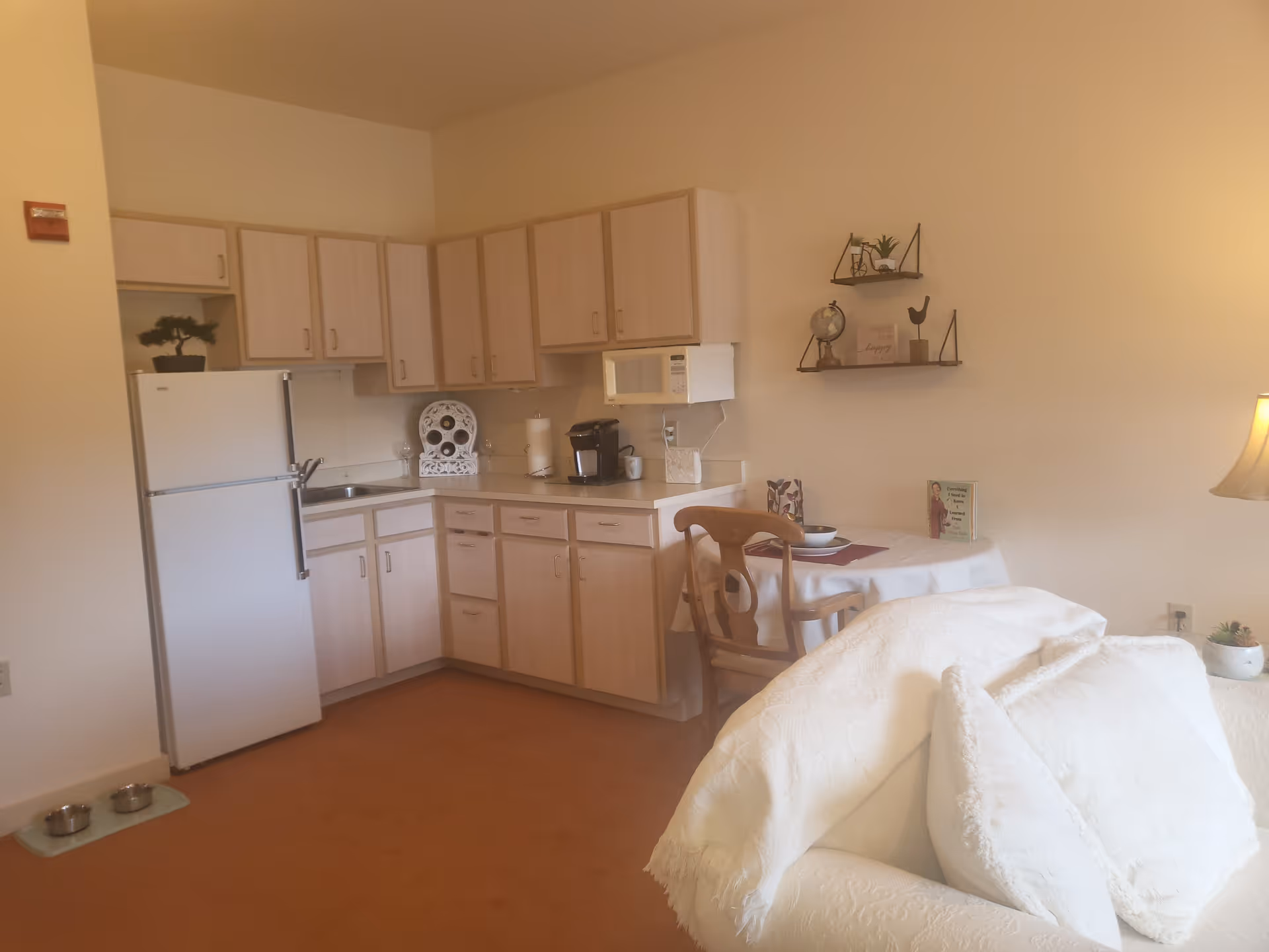 A cozy kitchen area with light wood cabinets, a white refrigerator, a microwave, a coffee maker, and a small sink. Adjacent to the kitchen is a small round dining table with two wooden chairs, set with a bowl and placemat. A white couch with white pillows is partially visible in the foreground, and a lamp and small plant are on a side table. Two small shelves with decorative items are mounted on the wall above the dining table.