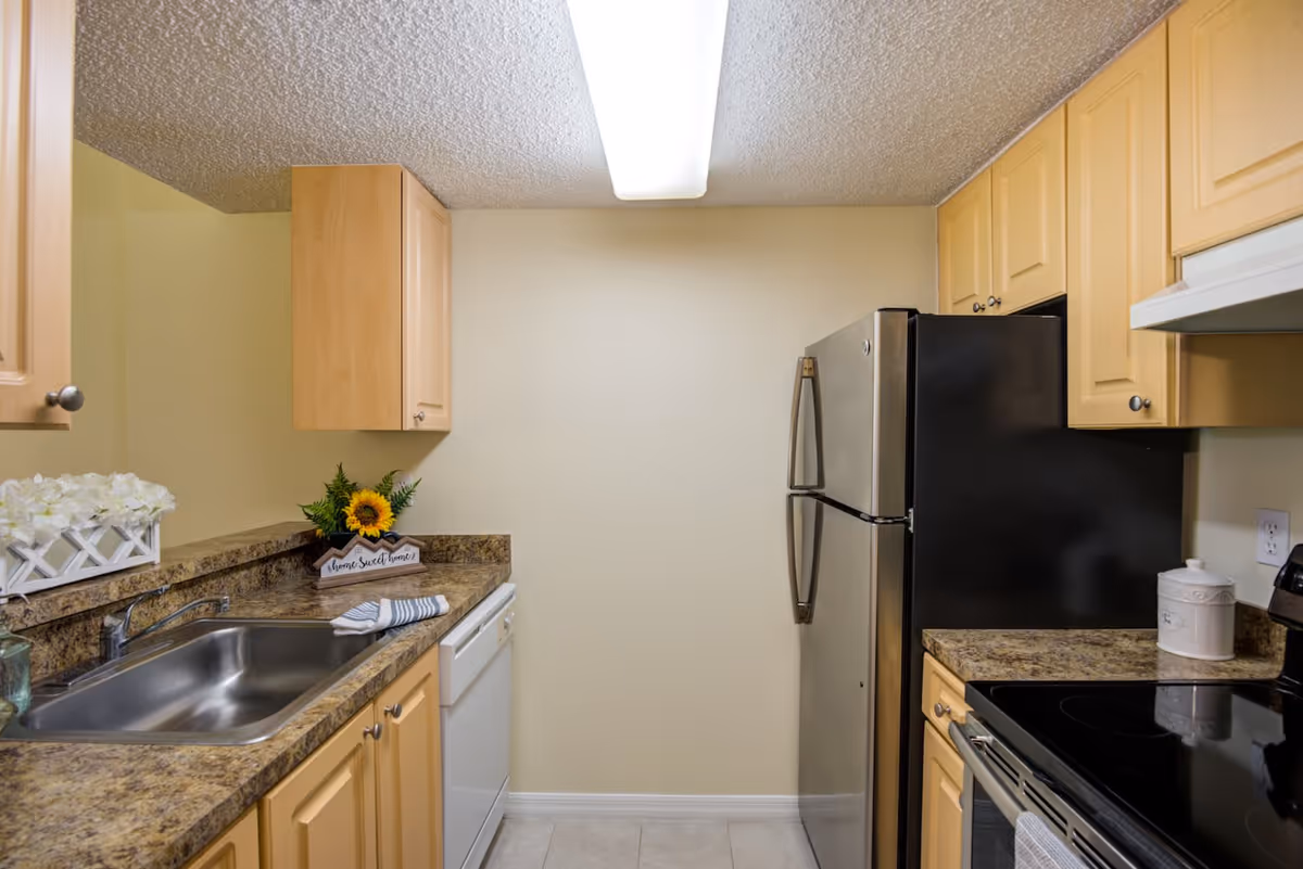 A compact kitchen with light wood cabinets, a stainless steel refrigerator, a black electric stove, a white dishwasher, and a stainless steel sink. The countertops are a brown granite pattern, and there is a small decorative arrangement with a sunflower and a sign that says 'home sweet home'. The walls are painted light beige and the ceiling has a textured finish with a fluorescent light fixture.