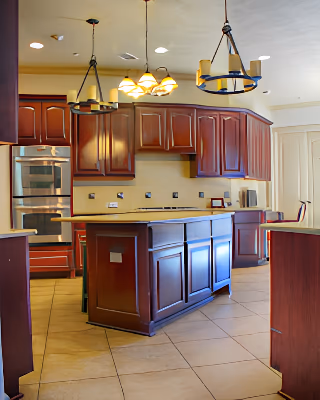 Spacious kitchen with dark wood cabinets, a large central island, tiled floor, and modern hanging light fixtures. Stainless steel double oven is visible on the left side.