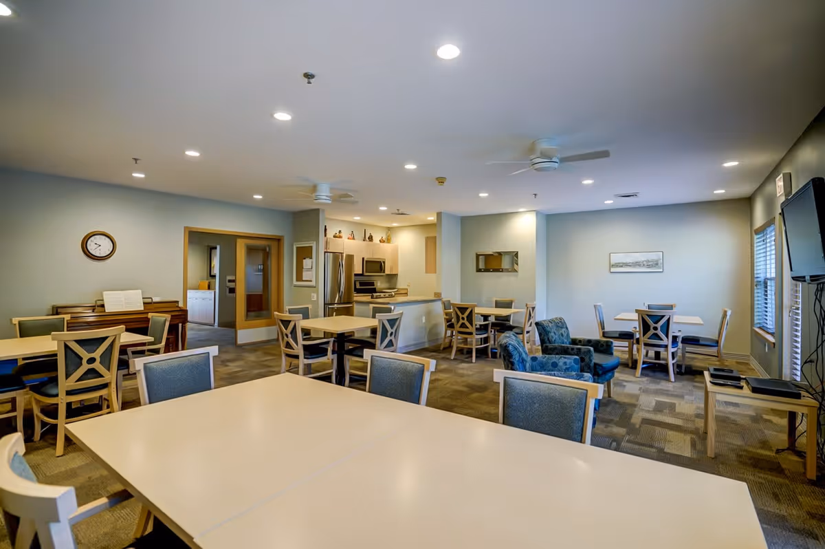 A spacious common area in a senior living facility featuring multiple tables and chairs arranged for dining or socializing. The room includes a small kitchen area with stainless steel appliances, a piano against one wall, a wall-mounted TV, and comfortable armchairs. The space is well-lit with recessed ceiling lights and ceiling fans, and has windows allowing natural light.