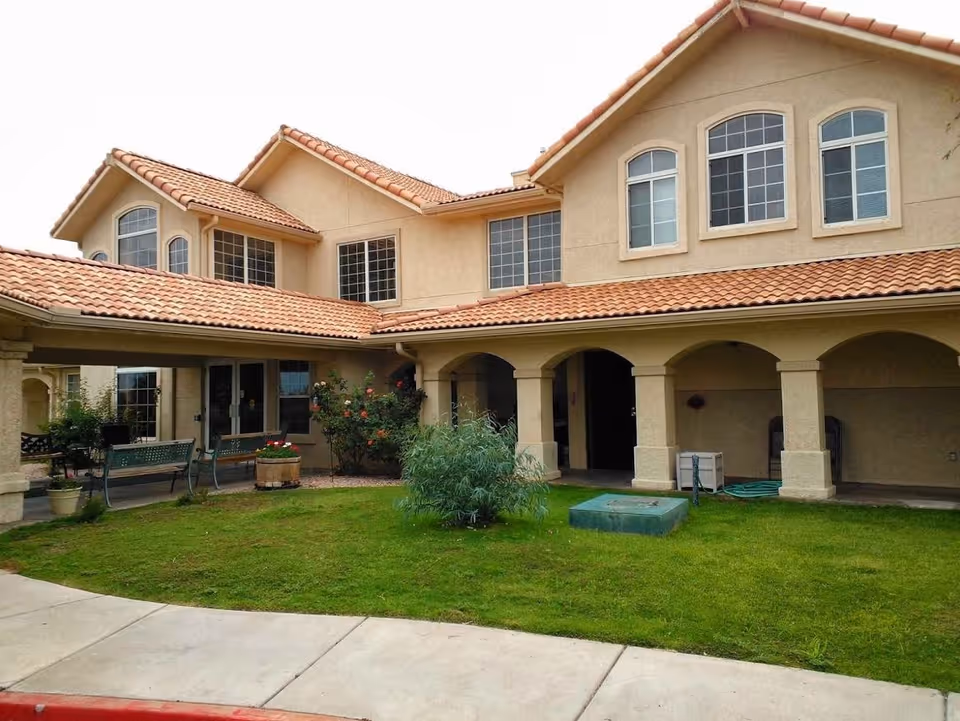 Exterior view of a two-story building with beige stucco walls and a red tile roof. The building has multiple windows and an arched covered walkway. There is a green lawn with some bushes and potted plants in front of the building, along with benches and a concrete sidewalk.
