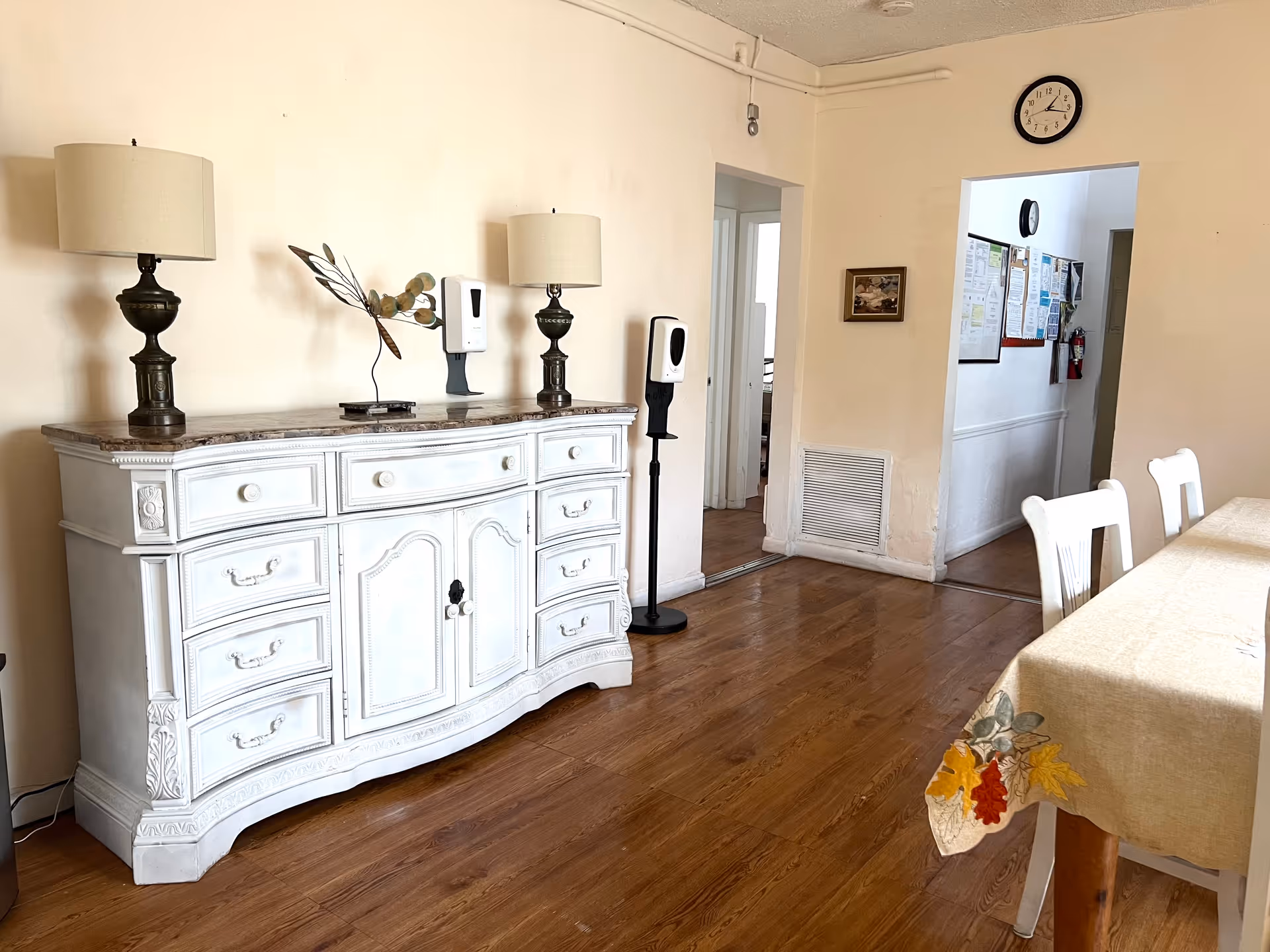 Dining room with a white ornate sideboard topped by two lamps and decor, hand-sanitizer dispensers, wooden floor, and a table with chairs.
