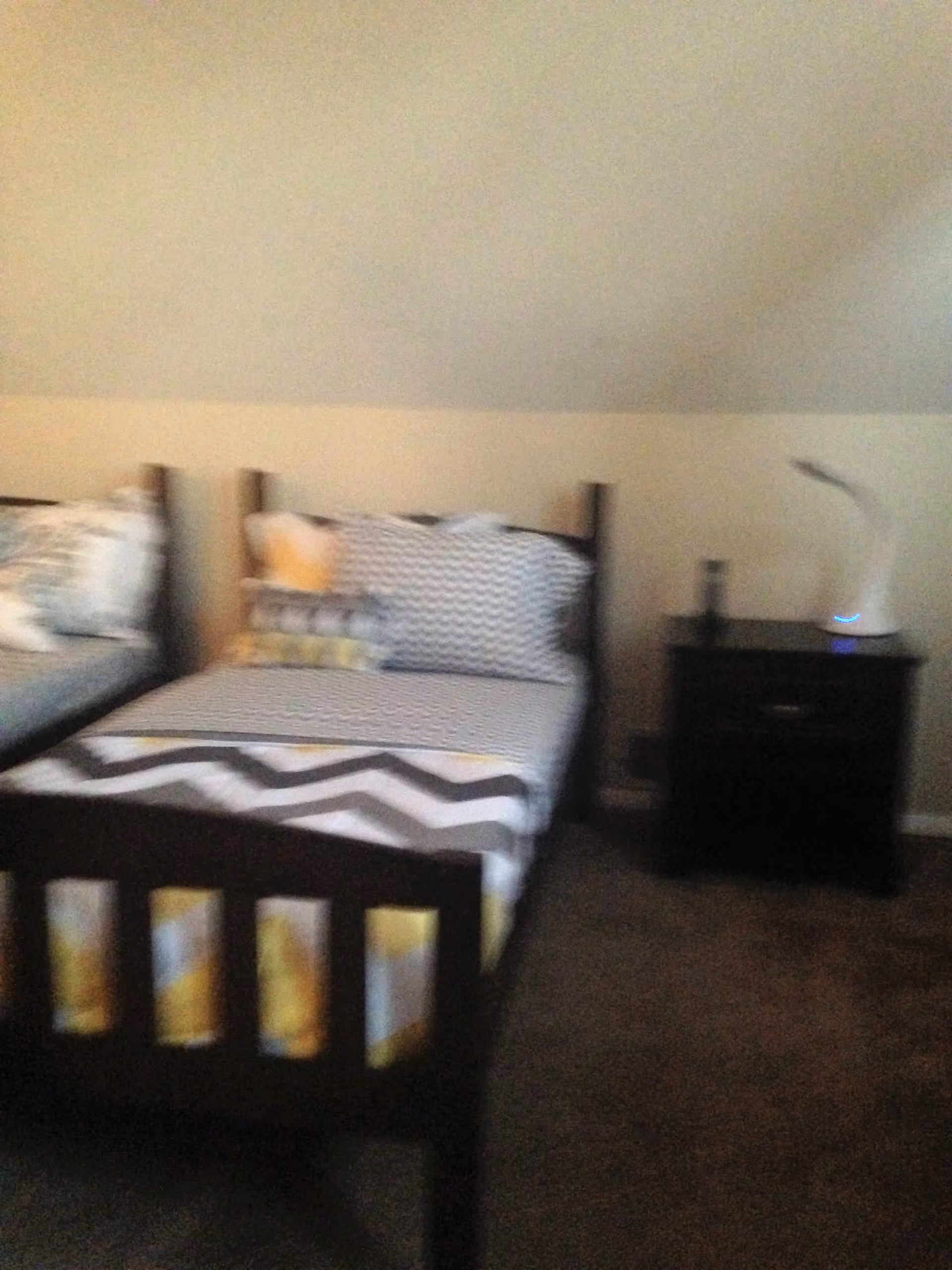 A bedroom with two single beds featuring patterned bedding in shades of gray, white, and yellow. There is a dark wooden nightstand between the beds with a white lamp on top. The walls are light-colored and the floor is carpeted.