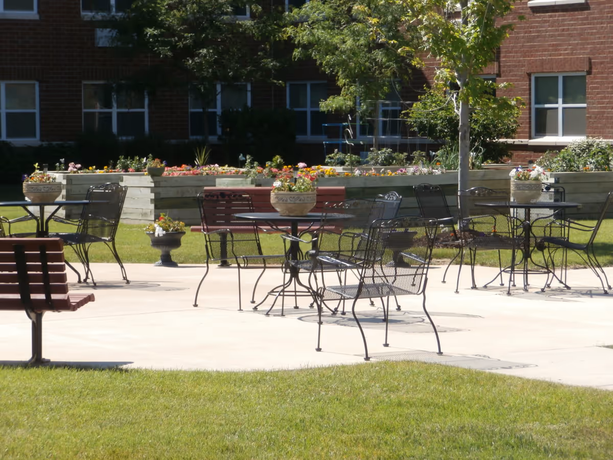 Outdoor patio area with metal tables and chairs, wooden benches, flower pots on tables, and raised garden beds with colorful flowers in front of a brick building.