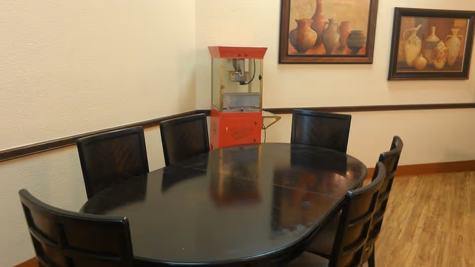A dining area with a black oval table surrounded by six black chairs. In the corner, there is a red popcorn machine. The walls are light-colored with two framed paintings of pottery hanging on them. The floor has a wood-like finish.
