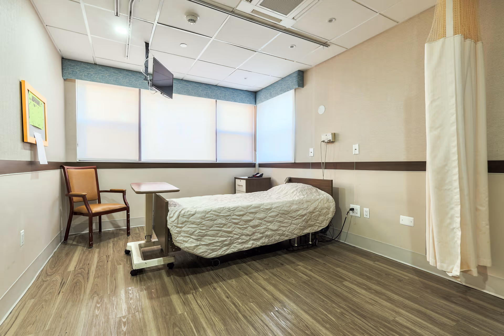 A small, clean hospital-style room with a single bed covered in a white quilted blanket. The room has wood-patterned flooring, a wooden chair, a small overbed table, a wall-mounted TV, and a privacy curtain on the right side. The walls are beige with a dark brown horizontal stripe, and there are large windows with blinds partially closed.