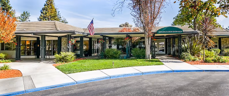 Front entrance of a single-story rehabilitation facility with a covered walkway, an American flag, and landscaped lawn.