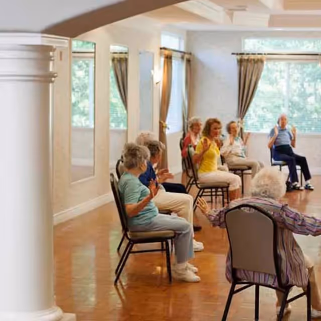 A group of elderly people sitting on chairs arranged in a spacious room with large windows and curtains, participating in a seated exercise or stretching activity.