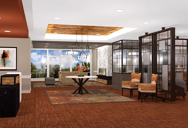A spacious and well-lit senior living facility common area with a large window showing trees outside. The room features a central round table with a flower arrangement, two upholstered chairs with orange and beige patterns, a modern fireplace with stacked wood, and a ceiling with wooden paneling and recessed lighting. The floor is covered with a reddish-brown carpet and a decorative rug under the table. Glass partitions separate different seating areas.