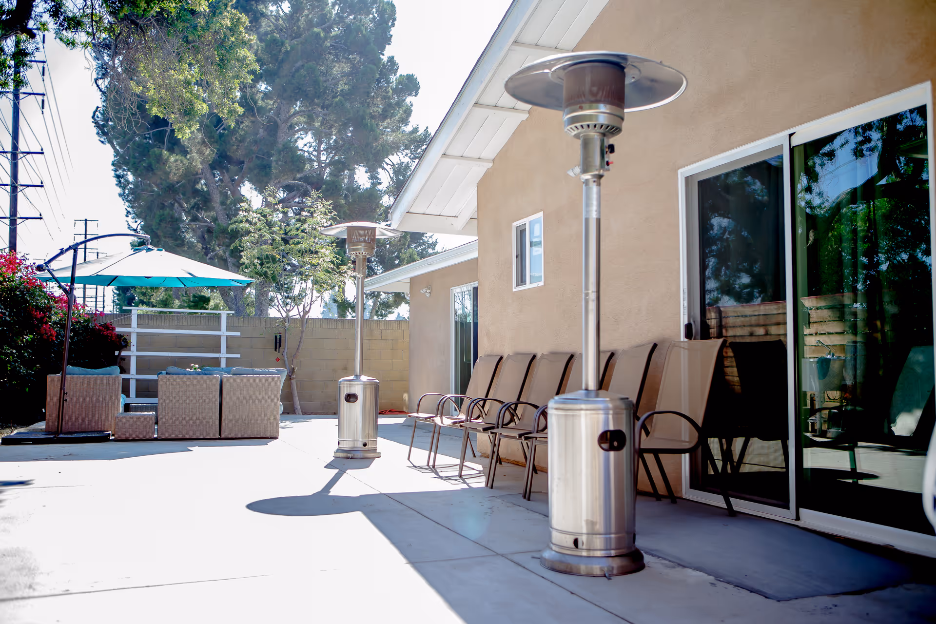Sunlit outdoor patio with seating, patio heaters, and an umbrella beside a sliding glass door.