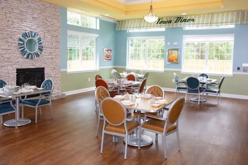 Bright dining room with multiple set tables and chairs, large windows, and a decorative stone fireplace.