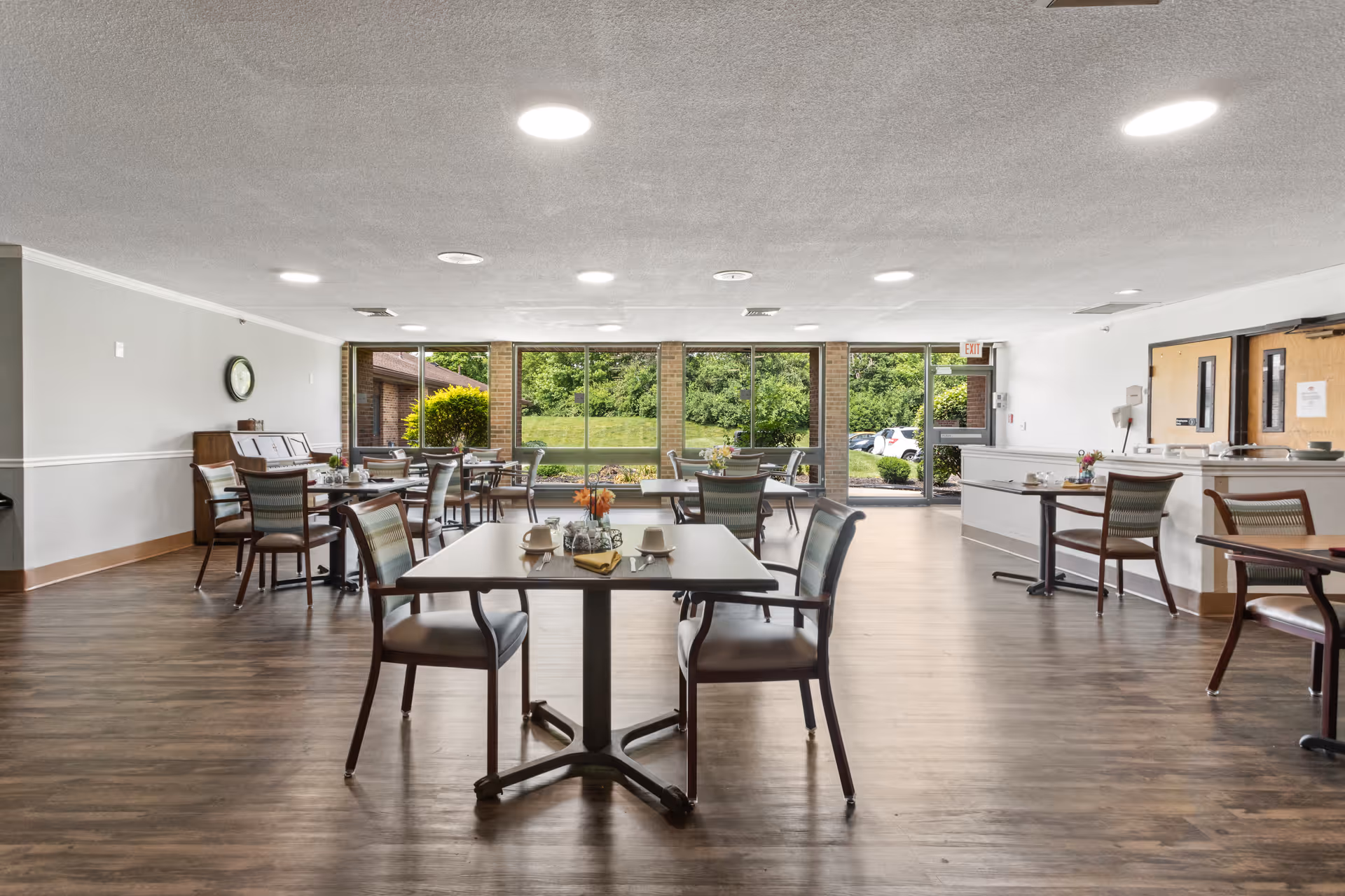 A spacious dining room with several tables and chairs arranged neatly. Each table has place settings including cups, plates, and napkins. Large windows along one wall provide a view of greenery and parked cars outside. The room has wood flooring, a piano against one wall, and bright ceiling lights.