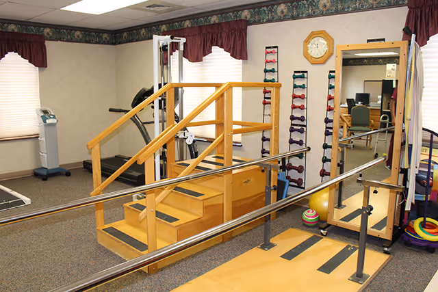 Interior view of a senior living facility exercise room featuring a wooden staircase with handrails for physical therapy, parallel bars for walking support, a large mirror, colorful dumbbells on racks, exercise balls, and various fitness equipment. The room has carpeted floors, beige walls with a floral border near the ceiling, and windows with maroon valances.