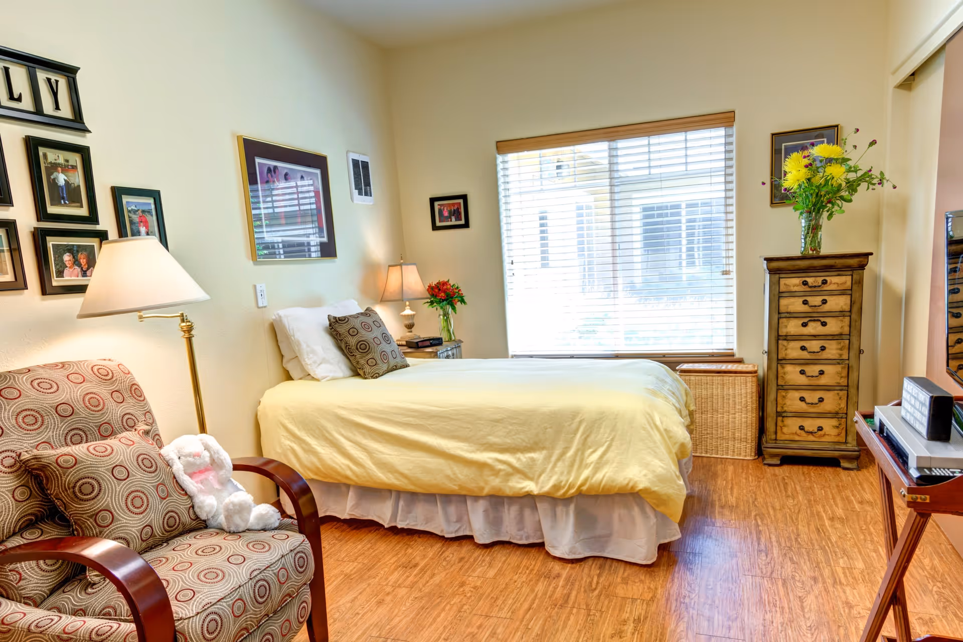 A cozy bedroom with a single bed covered in a yellow bedspread, a patterned armchair with a stuffed white bunny on it, a wooden chest of drawers with a vase of yellow flowers, framed pictures on the walls, and a large window with blinds letting in natural light.