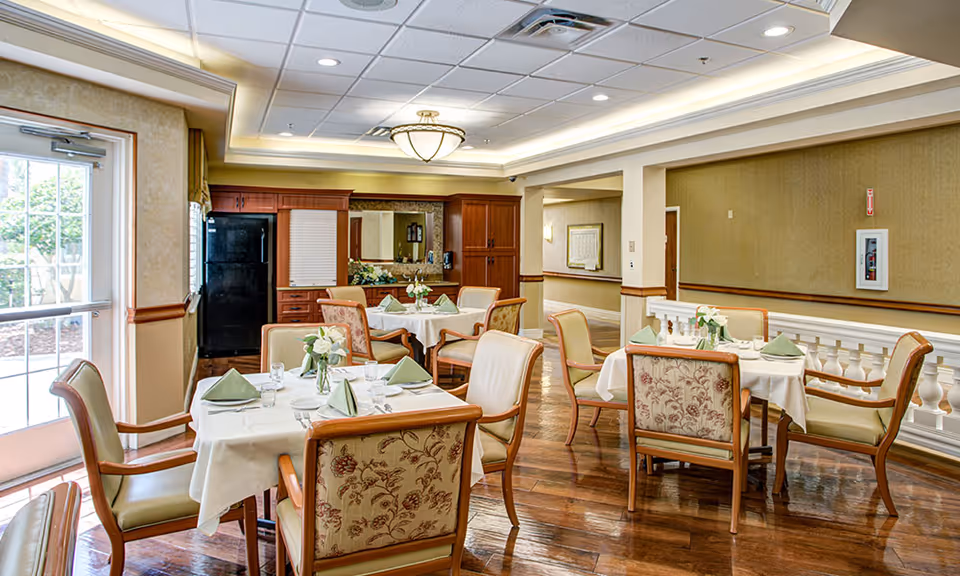 Well-lit dining room with several set tables, upholstered chairs, floral centerpieces, and wooden cabinetry in the background.