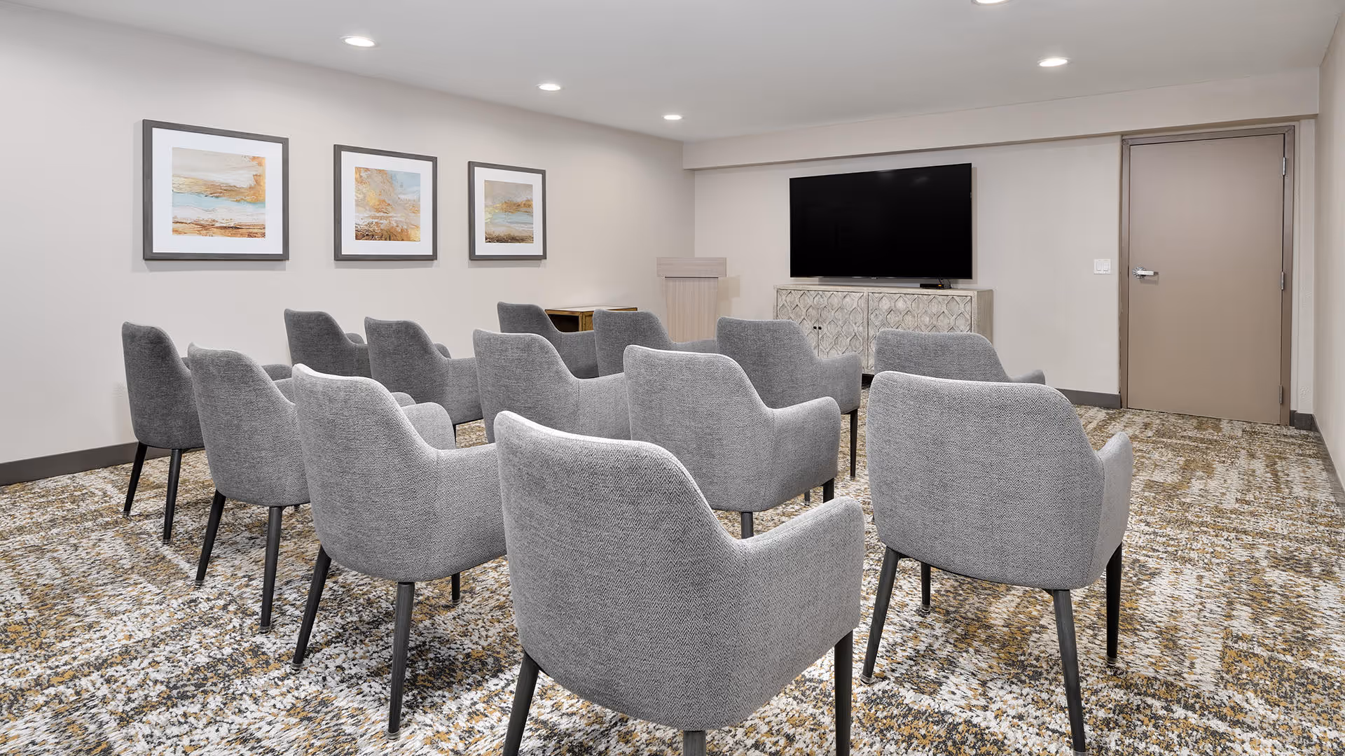 A small conference or media room with three rows of gray upholstered chairs facing a flat-screen TV mounted on the wall above a decorative cabinet. The room has a patterned carpet, neutral-colored walls, three framed abstract paintings, and recessed ceiling lights.