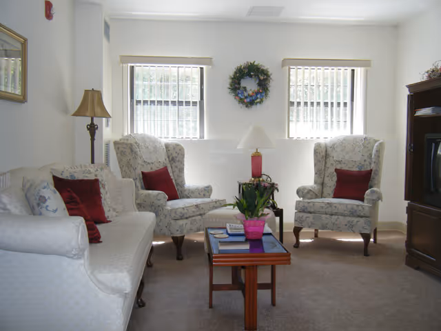 A cozy living room with a white sofa and two floral armchairs, each with a red cushion. A wooden coffee table with a pink potted plant and some magazines sits in the center. There is a floor lamp to the left and a table lamp on the coffee table. Two windows with vertical blinds and a decorative wreath are on the back wall. A wooden entertainment center with a TV is on the right side.