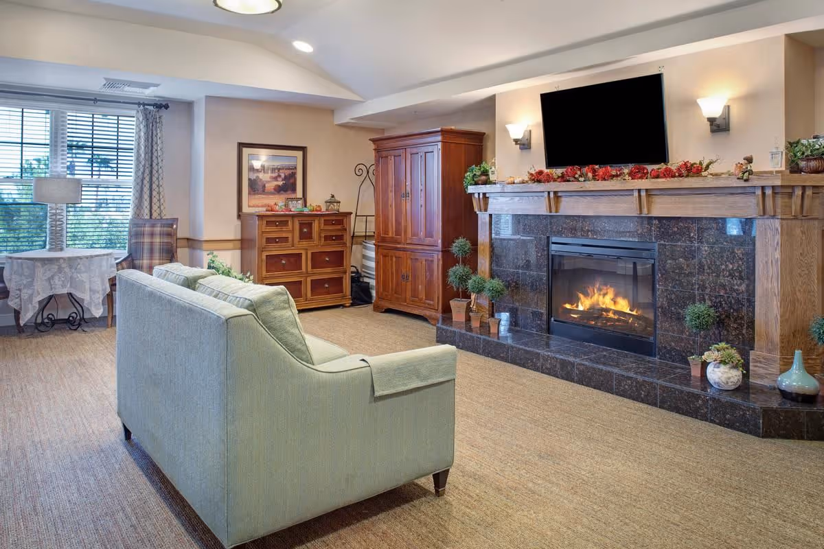 Cozy living room with a sofa facing a lit fireplace and wall-mounted TV, wooden cabinets, and a window.