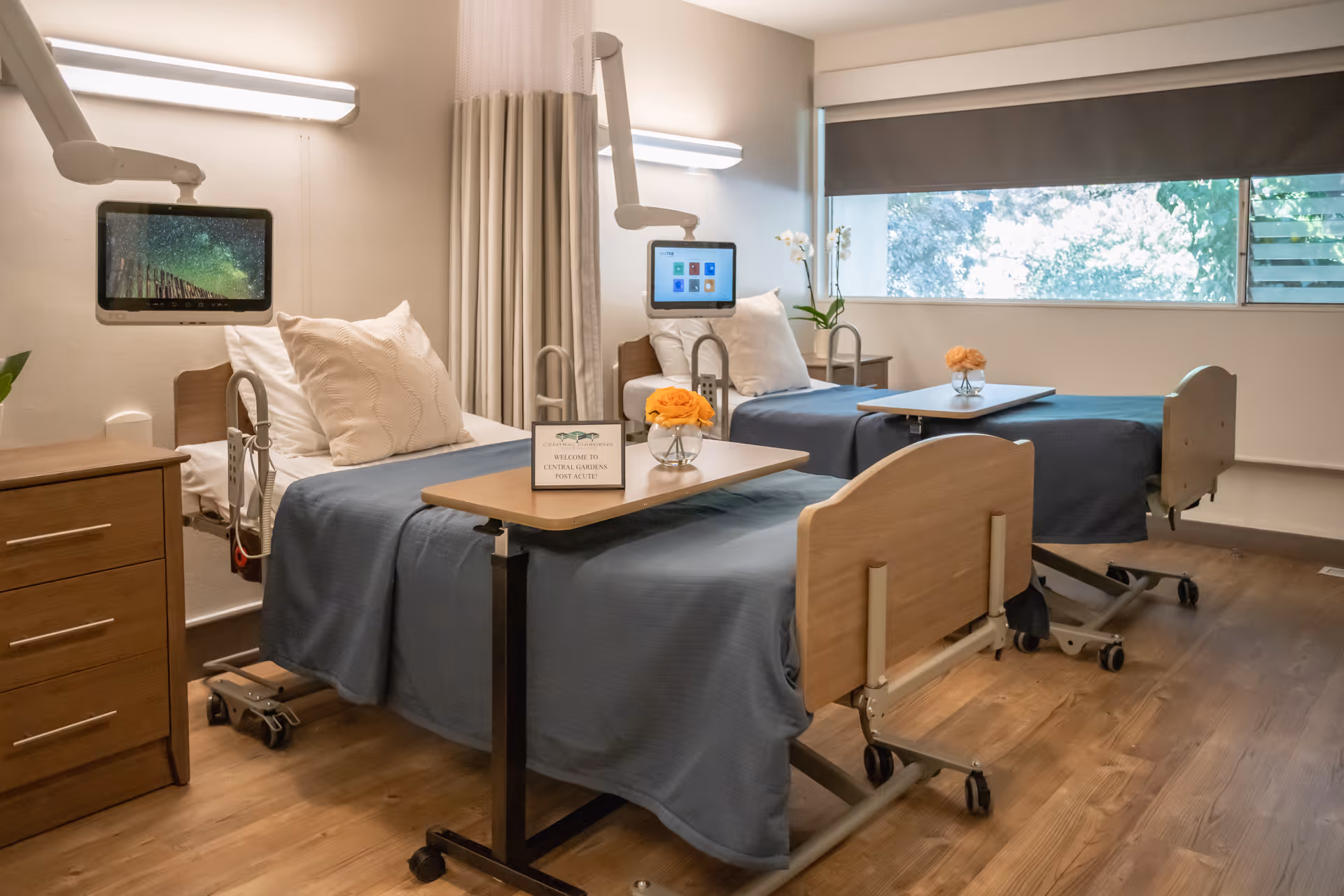 A bright and clean patient room with two hospital beds, each with a blue blanket and white pillows. Each bed has an adjustable overbed table with a small vase of orange flowers and a welcome sign. Mounted screens are positioned at the head of each bed, and there is a large window with a gray roller shade letting in natural light. The room has wooden flooring and a wooden bedside cabinet.