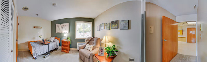 Sunlit senior resident bedroom with a bed, recliner, nightstands, wall art, and an open doorway to a hallway.