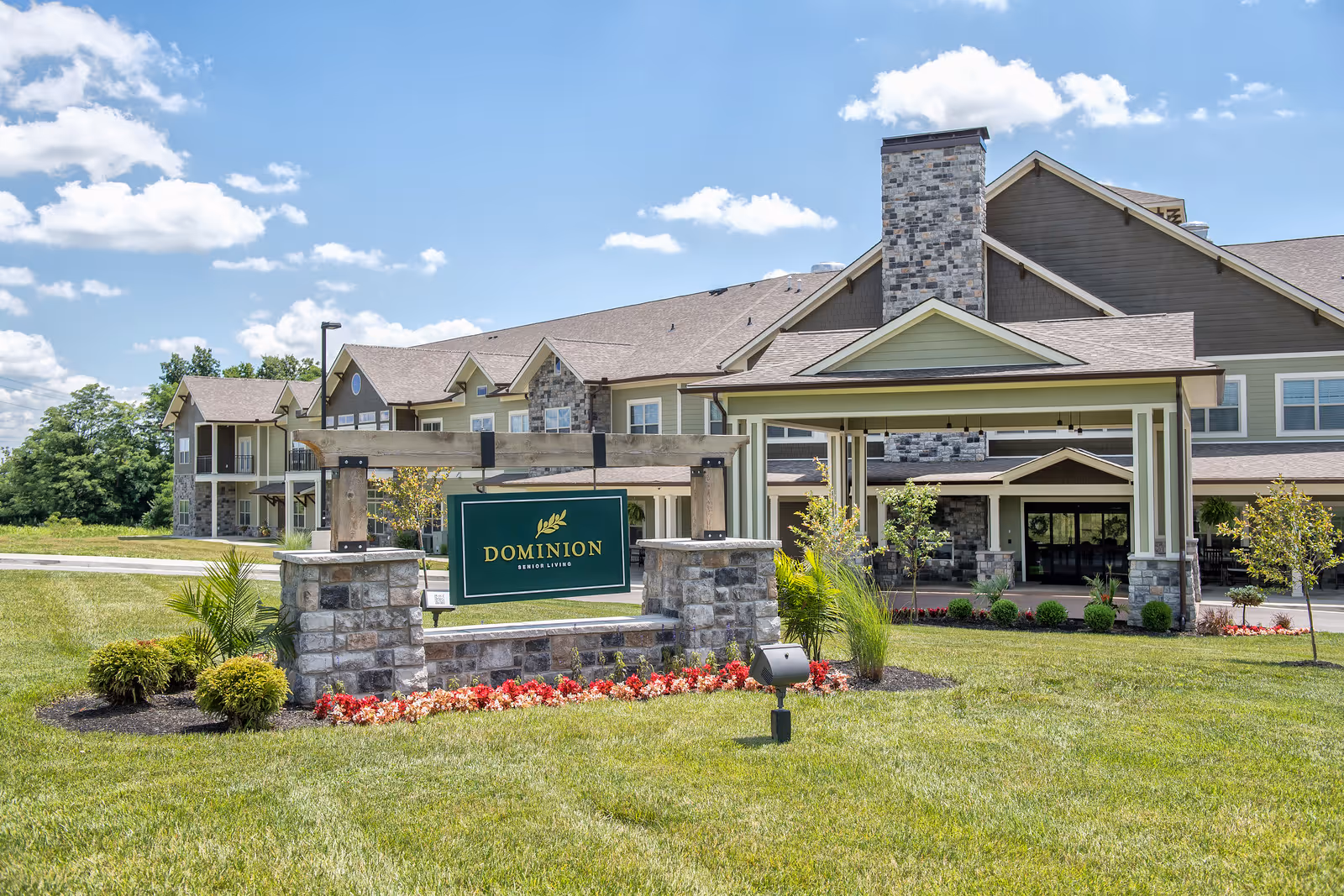 Front exterior of Dominion Senior Living building with entrance canopy, stone sign, and landscaped lawn on a sunny day.