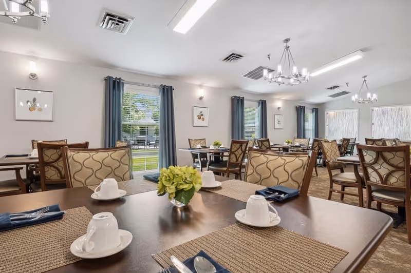 A bright dining room with multiple tables set with white cups, saucers, and silverware on woven placemats. The room has patterned chairs, large windows with blue curtains, and wall art. Outside the windows, greenery and outdoor seating are visible.