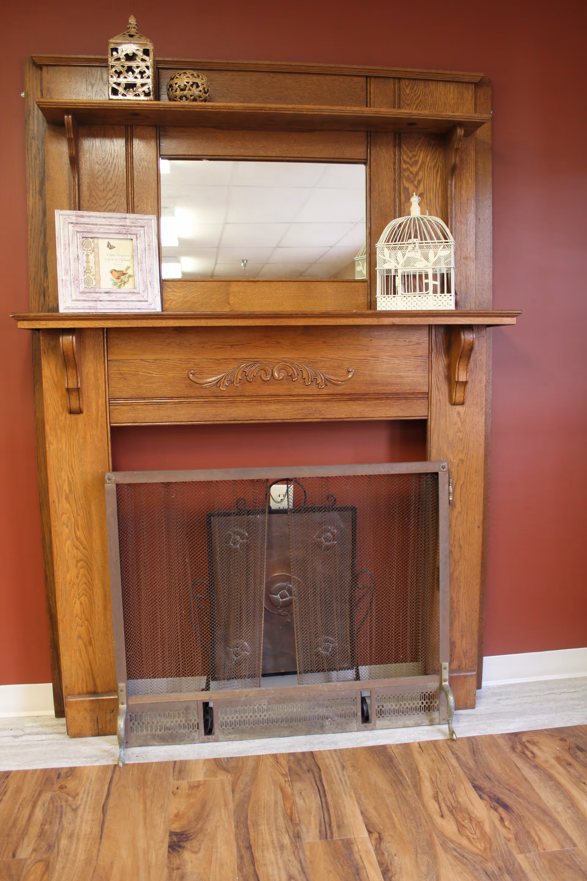 A wooden fireplace mantel with decorative carvings and a mesh screen in front. On top of the mantel are a framed picture, a decorative birdcage, and two ornamental lanterns. The wall behind is painted dark red and the floor has a wood pattern.
