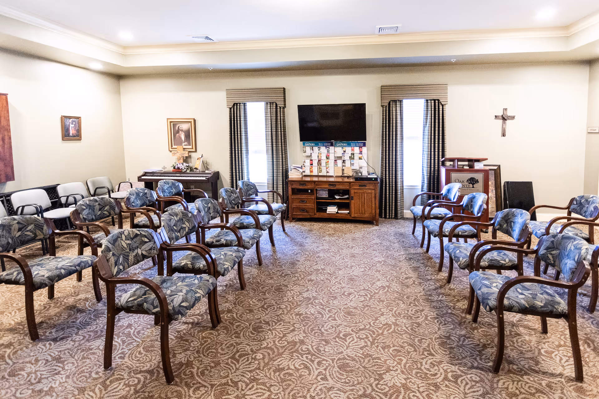 A meeting or activity room with multiple rows of upholstered chairs arranged facing a television mounted on the wall. The room has patterned carpet, two windows with curtains, a wooden cabinet under the TV, a piano, framed pictures on the walls, and a cross mounted on the right wall.