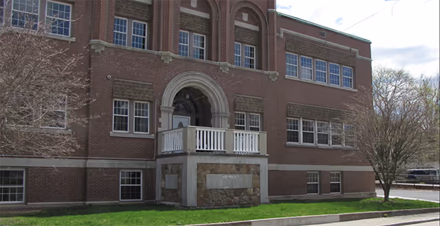 Brick three-story building front with an arched entrance, small balcony, and multiple windows above a grassy lawn.