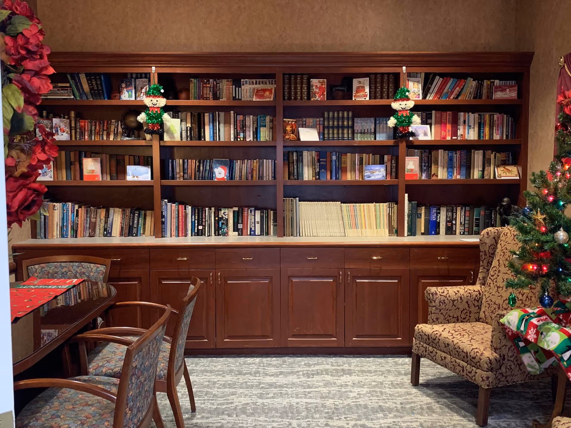 A cozy room with a large wooden bookshelf filled with books and holiday decorations including two small elf figures hanging on the shelves. To the left, there is a wooden table with chairs, and to the right, a patterned armchair next to a decorated Christmas tree with lights and ornaments. The room has a warm, inviting atmosphere with holiday decor.