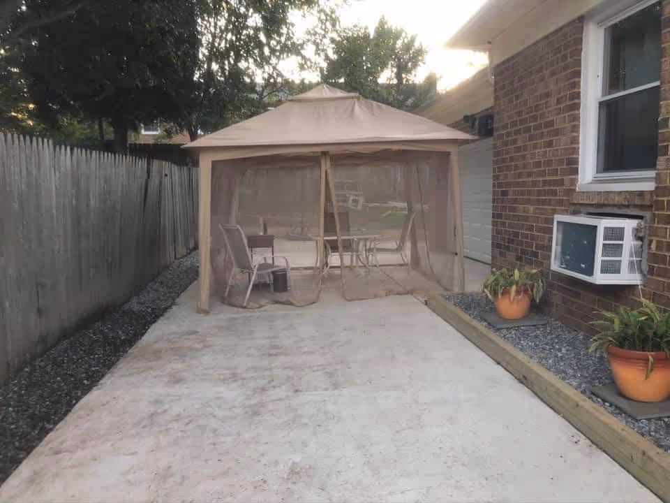 Outdoor patio area with a beige canopy tent enclosing a table and chairs. The patio is adjacent to a brick building with a window air conditioning unit and potted plants on a gravel bed. A wooden fence runs along the left side of the patio.