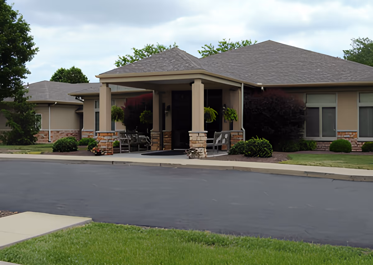 Exterior view of a single-story senior living facility building with a covered entrance supported by columns. The building has beige walls with stone accents and several windows. There are hanging plants and benches near the entrance, surrounded by well-maintained landscaping including bushes and grass. The sky is partly cloudy.