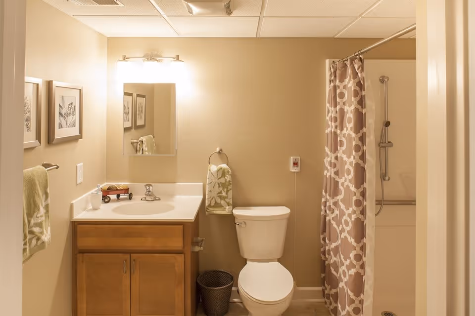 A bathroom with a wooden vanity cabinet, white sink, and a mirror above it. There are two green towels with white floral patterns hanging on towel racks. A white toilet is next to the vanity, and a shower with a beige and white patterned curtain is visible on the right. The walls are light beige, and there are two framed botanical prints on the left wall.