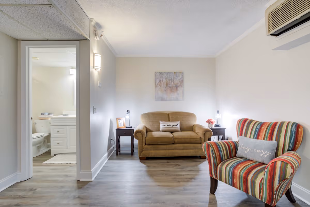 Small living room with a tan loveseat, colorful striped armchair, side tables with lamps, and a doorway opening to a bathroom.