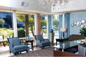 Bright sunlit common living room with blue walls, seating, rug, and large windows.