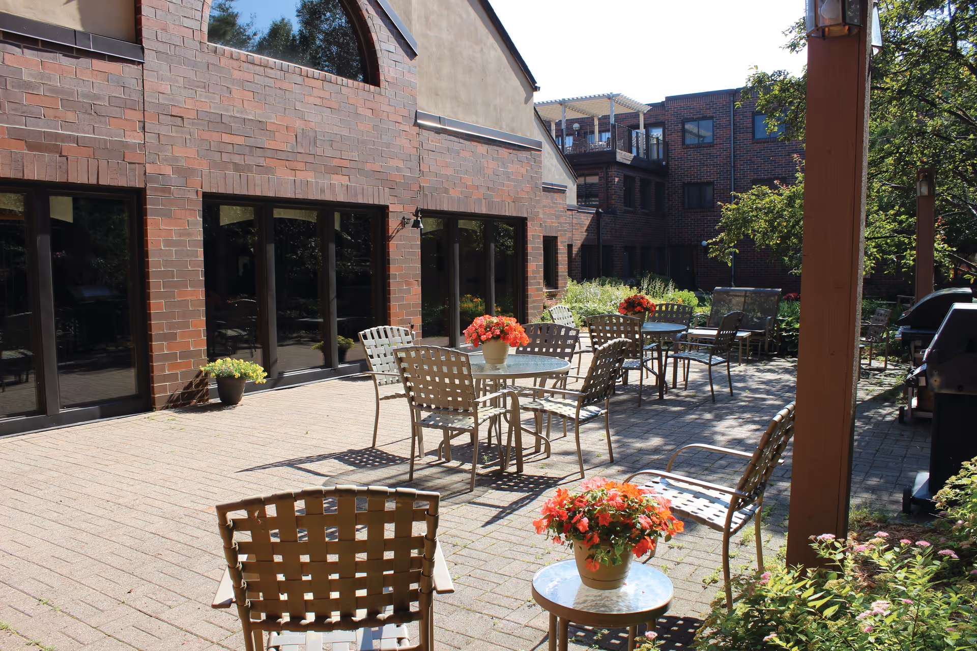 Sunlit brick courtyard patio with metal tables and chairs and potted flowers outside a brick senior living building.
